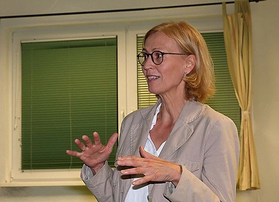 Eine Frau mit Brille und einer beigen Jacke spricht vor einem Fenster mit grünen Jalousien.