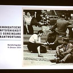 Ein Schwarz-Weiß-Foto zeigt eine Gruppe von Menschen auf Stufen. Die Bildunterschrift lautet 'Demokratische Zukunftsfähigkeit als gemeinsame Verantwortung'. Das Datum unten ist der 9. Oktober 2024.