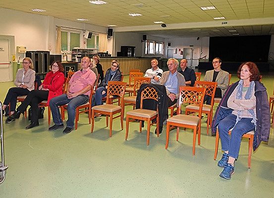 Eine Gruppe von Menschen sitzt in Stühlen in einem Raum mit einer Bühne und einem grünen Boden. Sie scheinen jemandem zuzuhören.
