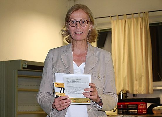 Eine Frau in einer hellbeigen Jacke hält einen Stapel Papiere in einem Raum mit gelben Vorhängen und einem Fenster.