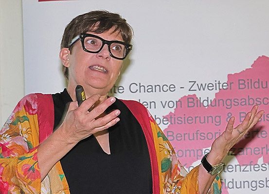 Eine Frau mit Brille spricht in ein Mikrofon und hält ein gelbes Werkzeug in der Hand, mit einem Schild im Hintergrund, das 'Zweite Chance - Zweite Bildung' besagt.