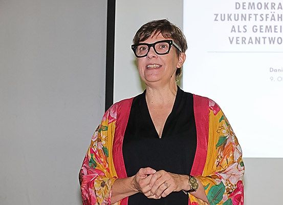 Eine Frau mit kurzen Haaren und einer Brille steht vor einer Projektionsleinwand mit deutschem Text.