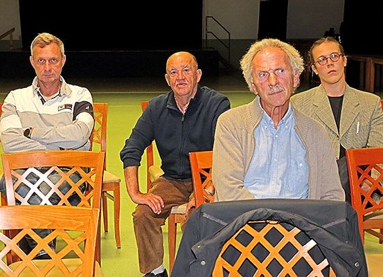 Vier Personen sitzen in Stühlen in einem Innenraum, wahrscheinlich einem Studio oder Theater. Jede Person hat einen einzigartigen Gesichtsausdruck, und eine trägt eine Brille. Ein Mantel hängt über der Rückenlehne eines Stuhls vor ihnen. Dahinter befindet sich eine Treppe und eine Wand.