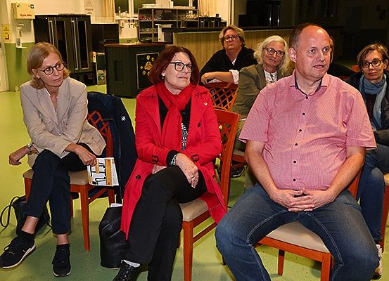 Eine Gruppe Erwachsener sitzt in Stühlen in einem Raum. Eine Frau in einem roten Mantel hält ein Buch. Ein Mann in einem rosa Hemd sitzt mit verschränkten Händen.