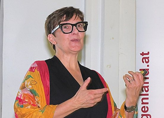 Eine Frau mit kurzen Haaren und einer Brille steht vor einer Wand und gestikuliert mit ihren Händen. Sie trägt eine farbenfrohe Jacke mit einem schwarzen Oberteil darunter. Die Wand hinter ihr hat ein rotes Schild mit dem Text 'genland.at'.