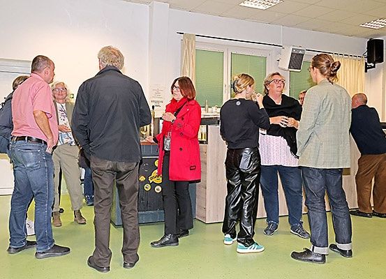 Eine Gruppe von Menschen steht und unterhält sich in einem Raum. Einige halten Tassen. Eine Frau in einem roten Mantel steht vor einer Theke.