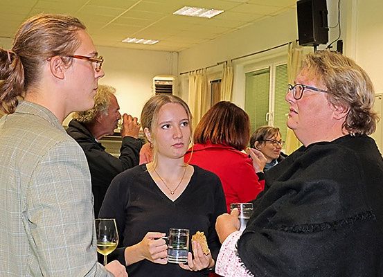 Eine Gruppe von Menschen in einem Raum, einige halten Gläser und andere unterhalten sich. Eine Frau in Schwarz hält ein Glas Wein.