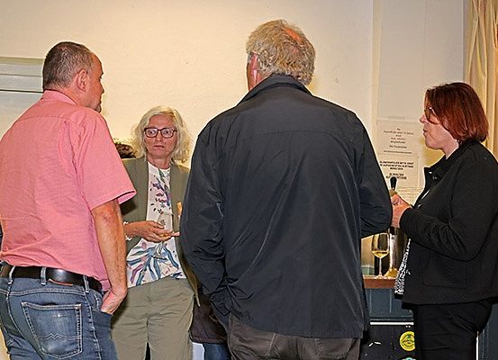 Vier Personen stehen in einem Raum, wobei sie sich gegenseitig anschauen und sich unterhalten. Die Frau auf der rechten Seite hält ein Weinglas. Hinter ihnen befindet sich eine Wand mit einem Schild und einer Flasche.
