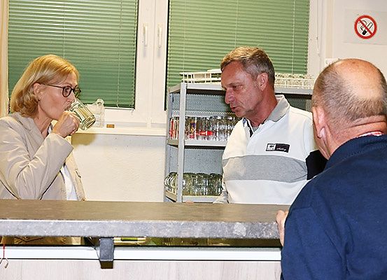 Drei Personen sind drinnen, in der Nähe einer Theke. Eine Frau trinkt aus einem Glas. Zwei Männer schauen sie an. Ein Regal steht hinter ihnen.