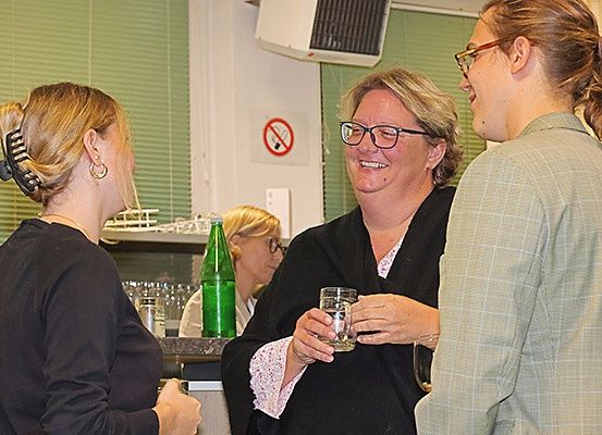 Drei Frauen stehen in einem Raum und unterhalten sich. Eine Frau hält ein Glas, während eine andere in die Kamera schaut. Eine grüne Flasche und ein Schild mit einem Rauchverbotssymbol sind im Hintergrund zu sehen.