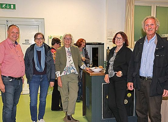 Fünf Menschen stehen lächelnd zusammen in einem Raum. Hinter ihnen ein Tisch mit Essen und Getränken. Eine Frau hält eine Schüssel und einen Teller. Im Hintergrund zwei Frauen, eine mit einem Glas. Eine Tür und Fenster sind sichtbar.