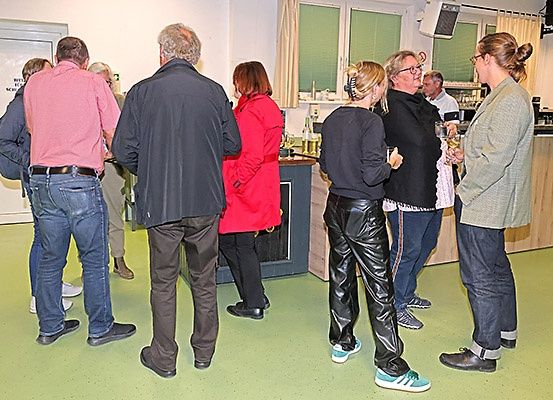 Eine Gruppe von Menschen versammelt sich in einem Raum mit grünem Boden und führt Gespräche, während sie Getränke in den Händen halten. Sie sind lässig gekleidet, einige tragen Brillen und Lederhosen. Der Raum hat Fenster mit Vorhängen und eine Tür auf der linken Seite.