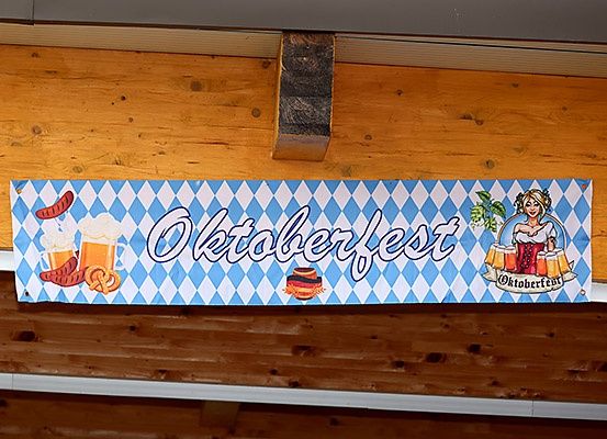 Ein blau-weiß kariertes Banner mit dem Wort Oktoberfest hängt an einer Holzwand. Es zeigt ein Bierglas, eine Brezel und Würste.