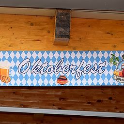 Ein blau-weiß kariertes Banner mit dem Wort Oktoberfest hängt an einer Holzwand. Es zeigt ein Bierglas, eine Brezel und Würste.