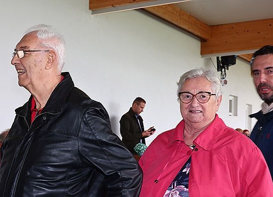 Drei Menschen stehen drinnen; ein älterer Mann in einer schwarzen Lederjacke, eine ältere Frau in einem rosa Mantel und ein Mann in einem blauen Mantel, wobei ein Mann im Hintergrund ein Handy benutzt.