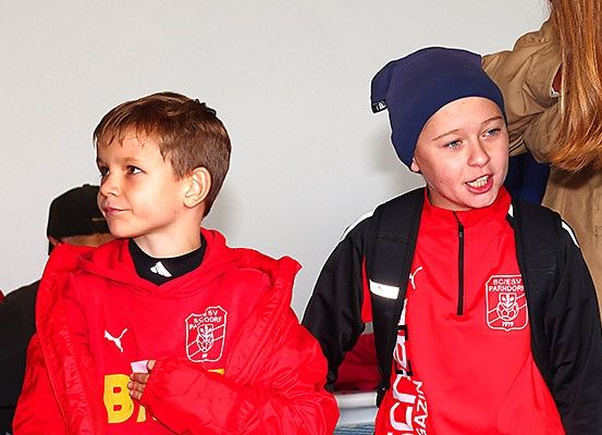 Zwei Jungen in roten Jacken, einer mit einem Logo, stehen zusammen, wahrscheinlich bei einer Sportveranstaltung. Hinter ihnen passt eine Frau mit langen Haaren ihre Jacke an.