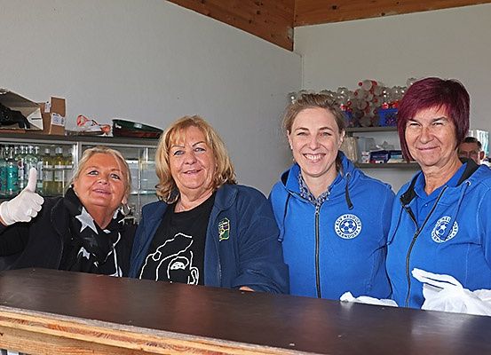 Vier Frauen stehen hinter einem Holztresen. Sie tragen alle blaue Jacken mit Logos. Eine von ihnen gibt einen Daumen hoch.