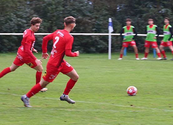 Zwei Fußballspieler in roten Trikots rennen auf einen Ball zu, der auf einem Feld liegt. Im Hintergrund befindet sich ein weißer Torpfosten. Andere Spieler in grünen Trikots stehen hinter dem Torpfosten.