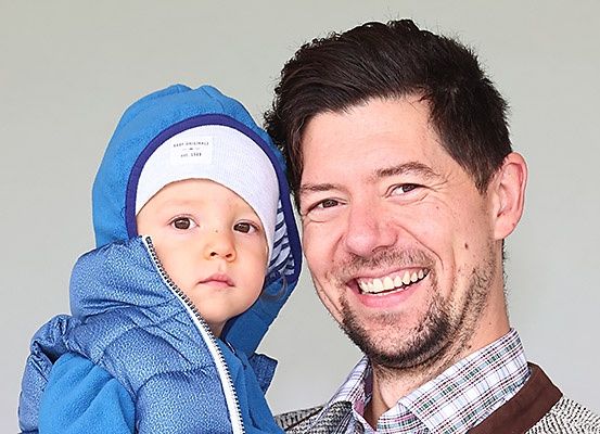 Ein Mann lächelt warmherzig, während er ein Baby in einer blauen Winterjacke und einer weißen Mütze hält, beide schauen in die Kamera.