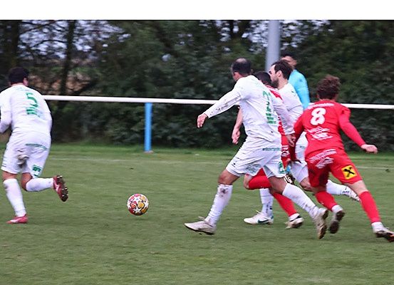 Fußballspieler in weißen und roten Trikots konkurrieren auf einem Feld mit einem Ball, Bäume im Hintergrund.
