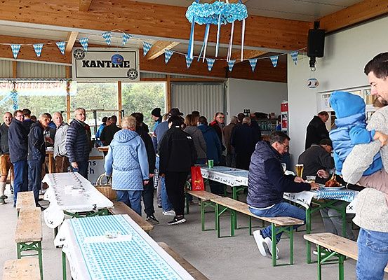 Eine Gruppe von Menschen steht in einer Schlange und sitzt auf Bänken in einer Kantine. Ein Mann hält ein Kind auf seiner Schulter. Tische haben blau-weiß karierte Tischdecken. Ein Lautsprecher hängt an der Wand.
