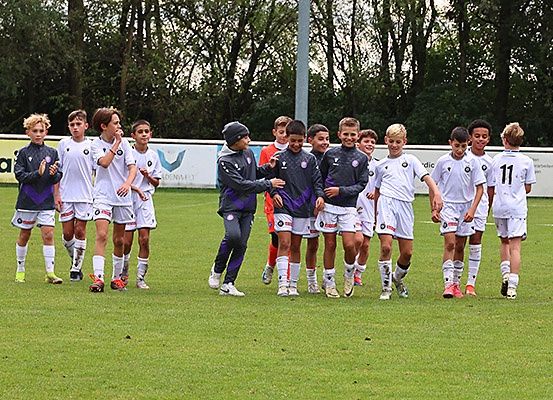 Eine Gruppe junger Fußballspieler in weißen Trikots läuft auf einem Rasenfeld, wahrscheinlich um einen Sieg zu feiern. Ein Mann in einem lila Jacke läuft vor ihnen.
