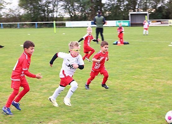 Eine Gruppe von Kindern in roten und weißen Trikots läuft auf einem Fußballfeld. Ein Fußball liegt vor ihnen auf dem Boden.