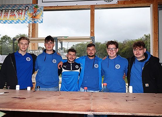 Eine Gruppe von Männern in blauen Jacken posiert für ein Foto vor einem Tisch. Auf dem Tisch befinden sich Flaschen und kleine Behälter. Hinter ihnen ist ein Fenster mit Blick auf Bäume und eine Fahne, die 'BIERFEST' steht.