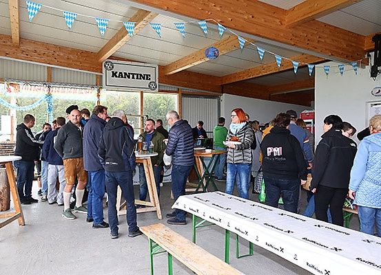 Eine Gruppe von Menschen steht in einer Schlange an einem Buffet in einem Restaurant. Einige tragen Jacken und Sneakers. Es gibt Tische mit weißen Tüchern und Bänken. Ein Schild an der Wand steht für 'KANTIN'.