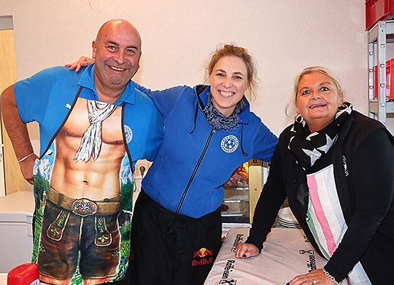 Drei Menschen stehen in einem Raum. Einer trägt ein T-Shirt mit einem Bild eines Mannes, ein anderer ein blaues Sweatshirt mit einem Logo und der dritte ein schwarzes Sweatshirt. Sie lächeln.