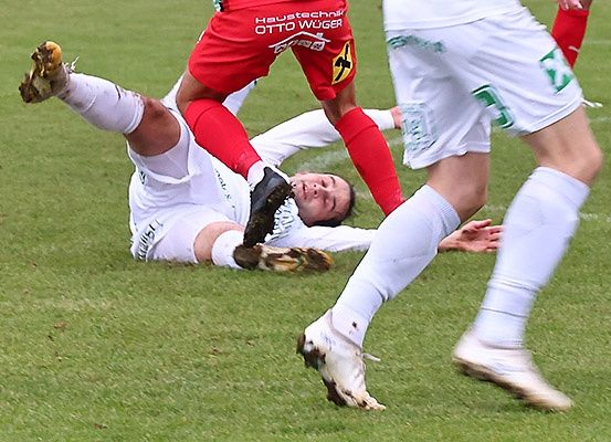 Zwei Fußballspieler in weißen und roten Trikots kollidieren auf einem Rasenfeld, einer liegt auf dem Boden und der andere steht mit einem Fuß auf dem Boden.