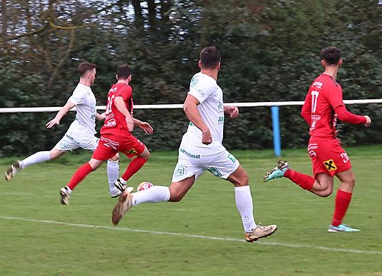 Vier Fußballspieler sind auf einem Feld und verfolgen einen Ball. Sie sind in der Luft, anscheinend im Wettstreit, um den Ball zuerst zu erreichen. Bäume und ein weißer Zaun sind im Hintergrund.