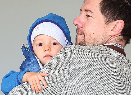 Ein Mann in einem Pullover hält ein Kind in einer blauen Jacke und einer weißen Mütze. Das Kind hat eine Hand auf der Schulter des Mannes.