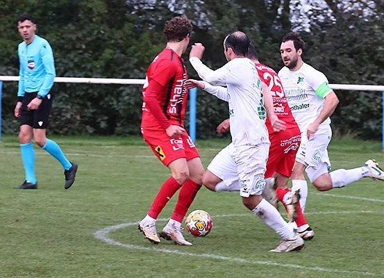 Vier Fußballspieler auf einem Feld. Zwei in Rot, zwei in Weiß. Ein Spieler tritt einen Ball, ein anderer versucht zu blocken. Ein Schiedsrichter steht am Rande.