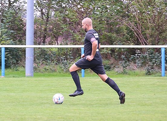 Ein Mann in einem schwarzen Trikot und Fußballschuhen tritt einen Fußball auf einem Rasenfeld mit einem Zaun dahinter.