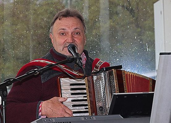 Ein Mann in einem burgunderroten Pullover spielt Akkordeon und singt in ein Mikrofon. Die Szene spielt sich draußen vor einem regnerischen Hintergrund ab.