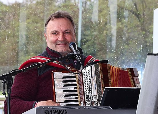 Ein Mann spielt ein Akkordeon an einem Mikrofon, mit einem Klavier vor ihm. Er lächelt und scheint zu spielen.