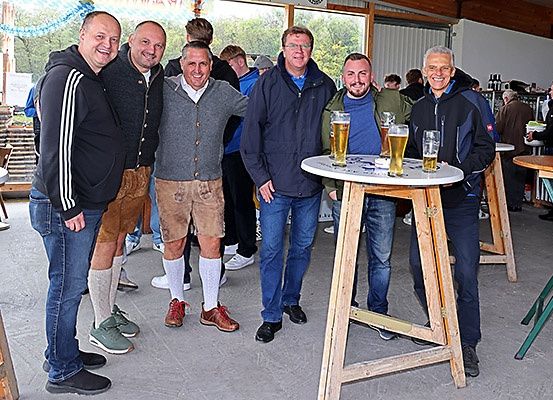 Fünf Männer, zwei mit Bierkrügen, stehen in einem Barbereich und lächeln. Hinter ihnen sind Tische und Stühle aufgestellt. Ein Mann trägt braune Hosen und ein anderer eine Jacke. Draußen sind mehr Leute und Bäume.