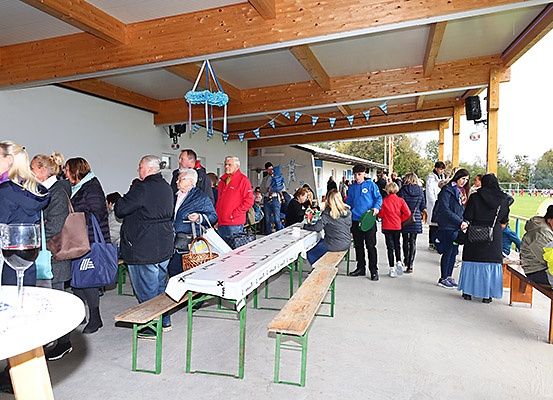 Menschen versammeln sich in einem Pavillon, einige sitzen auf Bänken und andere stehen. Lange Tische sind mit weißen Tüchern bedeckt. Weingläser stehen auf einem Tisch. Eine blaue Tasche ist auf der Bank. Ein Lautsprecher ist an der Wand.