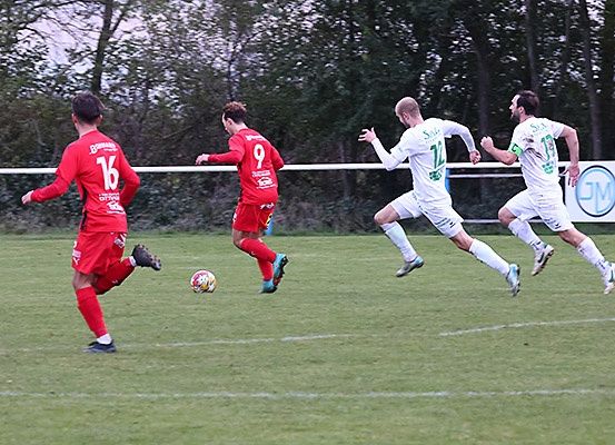 Ein Fußballspiel ist im Gange, zwei Spieler in Rot jagen den Ball, während zwei in Weiß verteidigen. Das Feld ist grün mit weißen Markierungen.