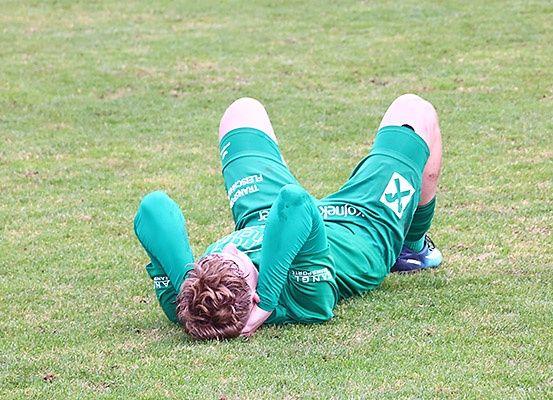 Ein Fußballspieler liegt auf dem Boden, möglicherweise verletzt, in einem grünen Trikot mit einem Kreuzlogo auf der Brust.