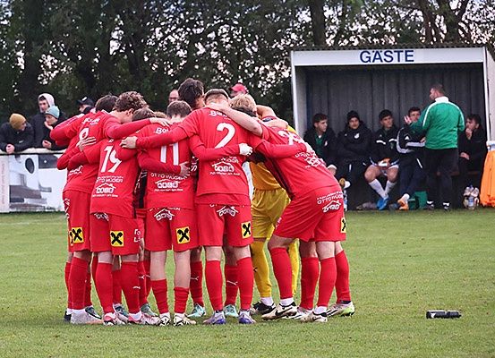 Eine Fußballmannschaft in roten Trikots ist auf dem Feld versammelt, mit Zuschauern im Hintergrund.