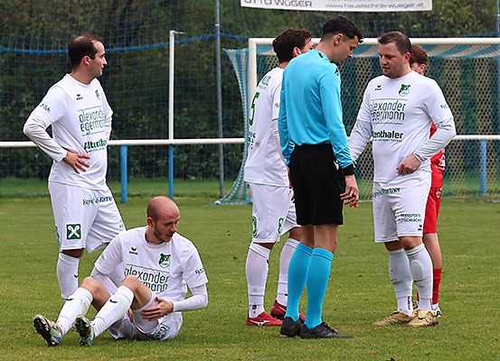Ein Fußballspiel ist unterbrochen; Spieler und Schiedsrichter sind versammelt, ein Spieler sitzt auf dem Boden, und eine Werbebanner im Hintergrund.