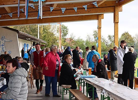 Menschen versammeln sich in einem offenen Essbereich unter einem Pavillon mit blau-weißen Wimpelketten, einige sitzen an Tischen mit Essen und Getränken, andere stehen und unterhalten sich, alle warm gekleidet.