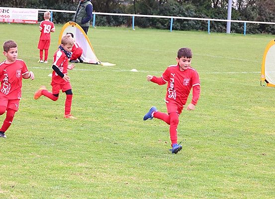 Mehrere junge Jungen rennen auf einem Fußballfeld in roten Trikots und trainieren für ein Spiel. Im Hintergrund sind ein Tornetz und ein Zelt zu sehen.