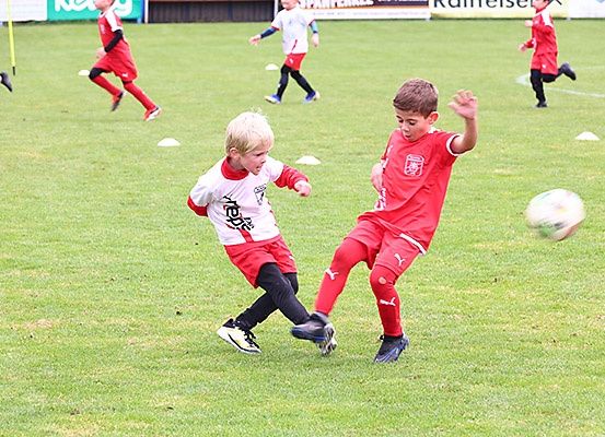 Kinder in roten Trikots spielen auf einem Fußballfeld. Zwei Jungen jagen dem Ball hinterher, einer in einem weißen Trikot.