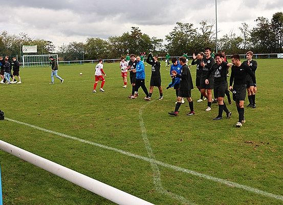 Eine Fußballmannschaft trainiert auf einem Feld, mit Spielern in verschiedenen Trikots und einem Ball in der Nähe. Das Feld hat weiße Markierungen und ist von Bäumen und einem Zaun umgeben.