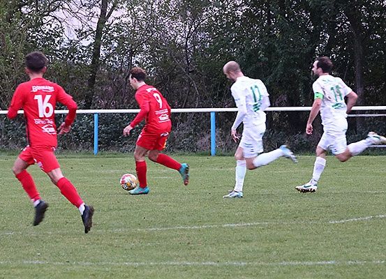 Vier Fußballspieler in roten und weißen Trikots spielen auf einem Rasenfeld. Sie jagen dem Ball hinterher, wobei Spieler in weißen Trikots zum Tor rennen.