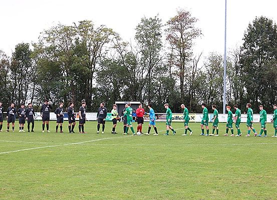 Zwei Fußballmannschaften stehen auf einem Feld und tragen Uniformen und Schuhe. Einige Spieler schütteln Hände. Im Hintergrund sind Bäume zu sehen.
