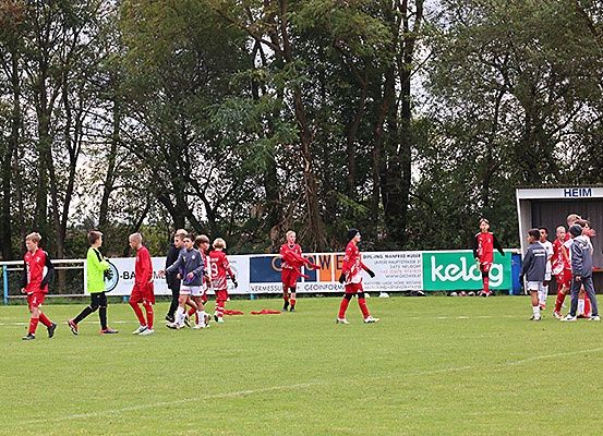 Ein Fußballspiel in vollem Gange, bei dem die Spieler rote Trikots tragen und ein Schiedsrichter in Grün das Spielfeld überquert. Im Hintergrund sind Bäume und eine Werbetafel zu sehen.